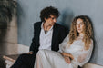 A man in a dark suit and a woman clad in Noémiah's Charlotte Dress — Bridal Edit, made of 100% silk organza, sit closely on a bench against a gray wall. The non-traditional bride, with curly hair and a serene expression, captivates the man's intent gaze. A plant decorates the background.