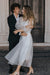 A non-traditional bride and her partner stand in front of a large wooden door, both smiling warmly. The man is in a black suit, while she wears a sheer Charlotte Dress from the Bridal Edit collection by Noémiah, crafted from 100% silk organza with transparent sleeves. They embrace lovingly on the cobblestone path.