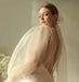 A woman in a sophisticated Charlotte Dress — Bridal Edit by Noémiah, crafted from 100% silk organza and featuring stylish puffed sleeves, looks back over her shoulder. She has a delicate veil tied in her hair and holds a bouquet of small white flowers, casting a serene glow that is ideal for non-traditional brides.