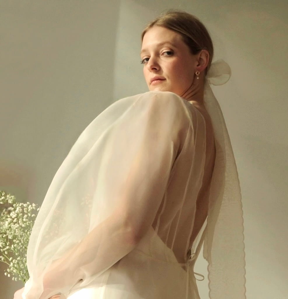 A woman in a sophisticated Charlotte Dress — Bridal Edit by Noémiah, crafted from 100% silk organza and featuring stylish puffed sleeves, looks back over her shoulder. She has a delicate veil tied in her hair and holds a bouquet of small white flowers, casting a serene glow that is ideal for non-traditional brides.