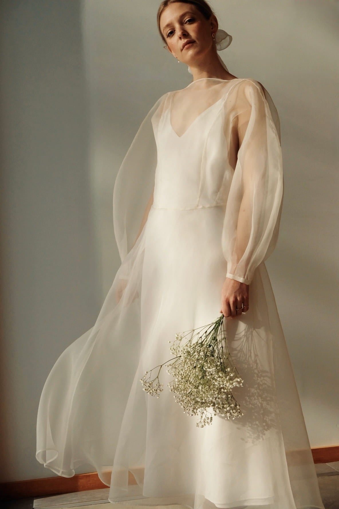 A person adorned in the Charlotte Dress — Bridal Edit by Noémiah stands in a softly lit room, holding a bouquet of white flowers. The gown's long, billowing sleeves and its 100% silk organza fabric create an elegant and serene appearance, perfect for non-traditional brides.