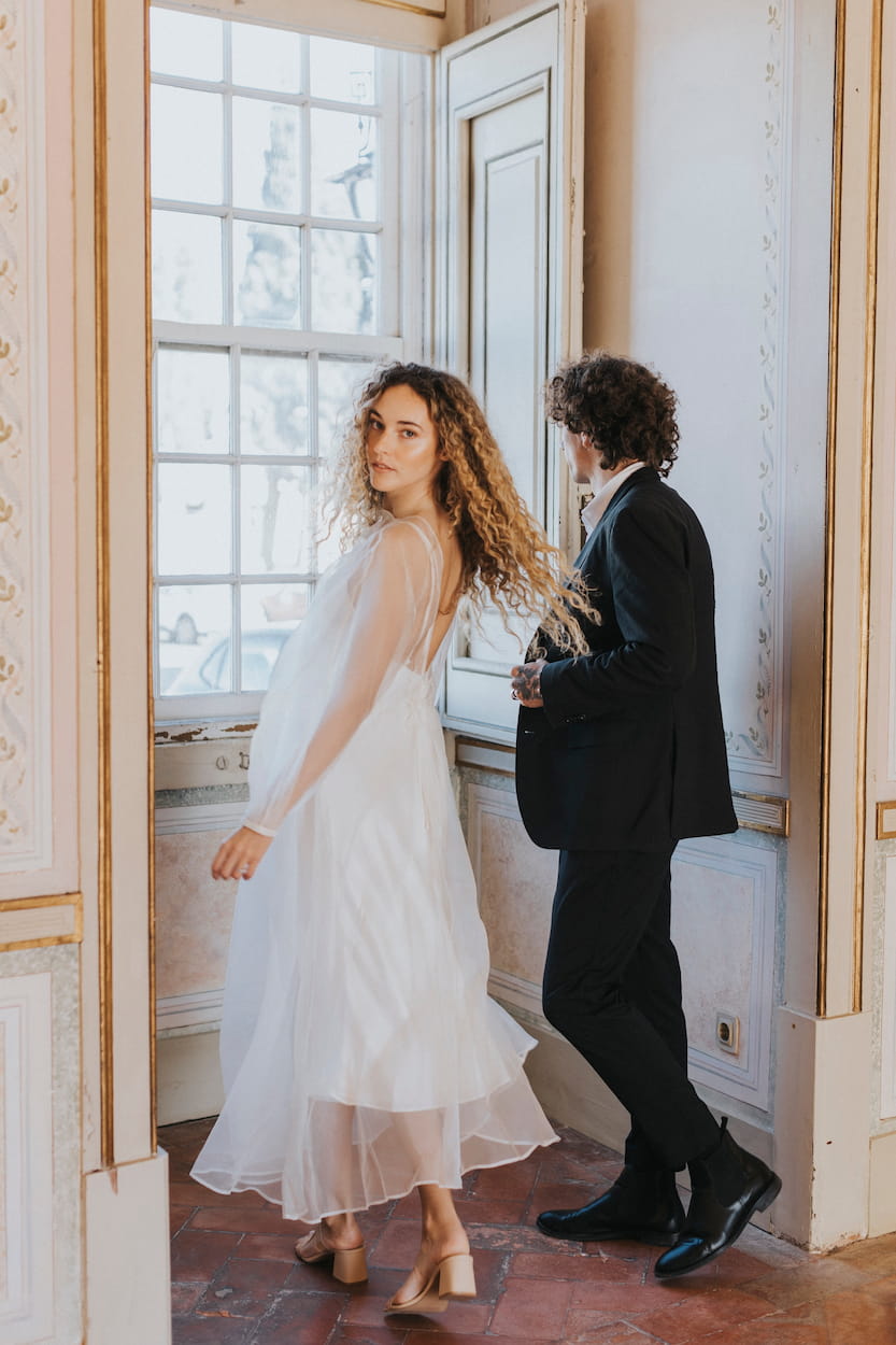 A woman wearing the Charlotte Dress from Noémiah's Bridal Edit, crafted from 100% silk organza, stands alongside a man in a black suit beside an ornate window in an elegantly decorated room. The non-traditional bride glances back over her shoulder as she walks, while the man gazes outside. Sunlight streams through the window.