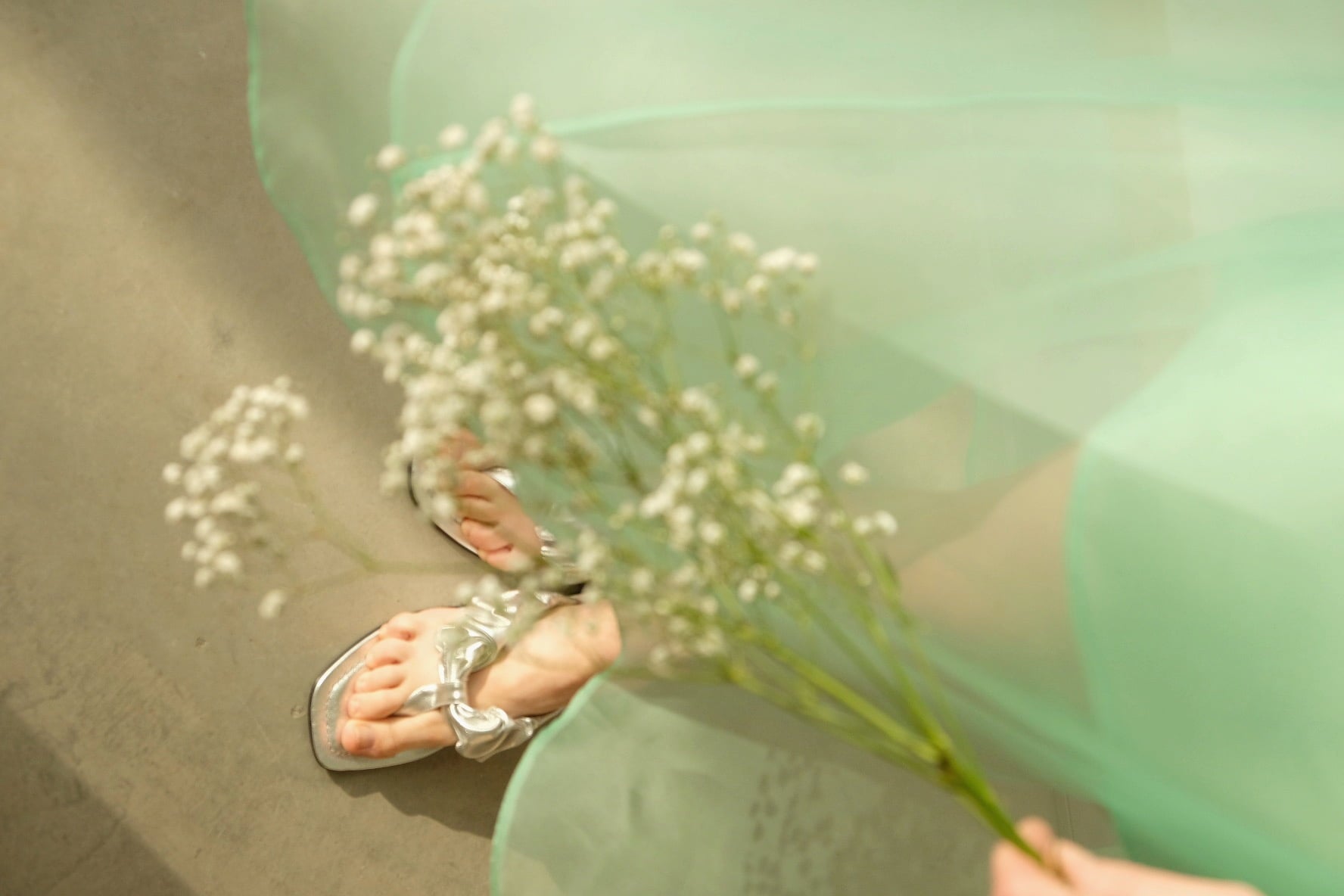 A person wearing transparent sandals holds a sprig of baby's breath flowers, their feet partially covered by the light green sheer fabric of the Charlotte Dress — Classic from Noémiah, made of 100% silk organza, creating a soft, ethereal look on a neutral background.