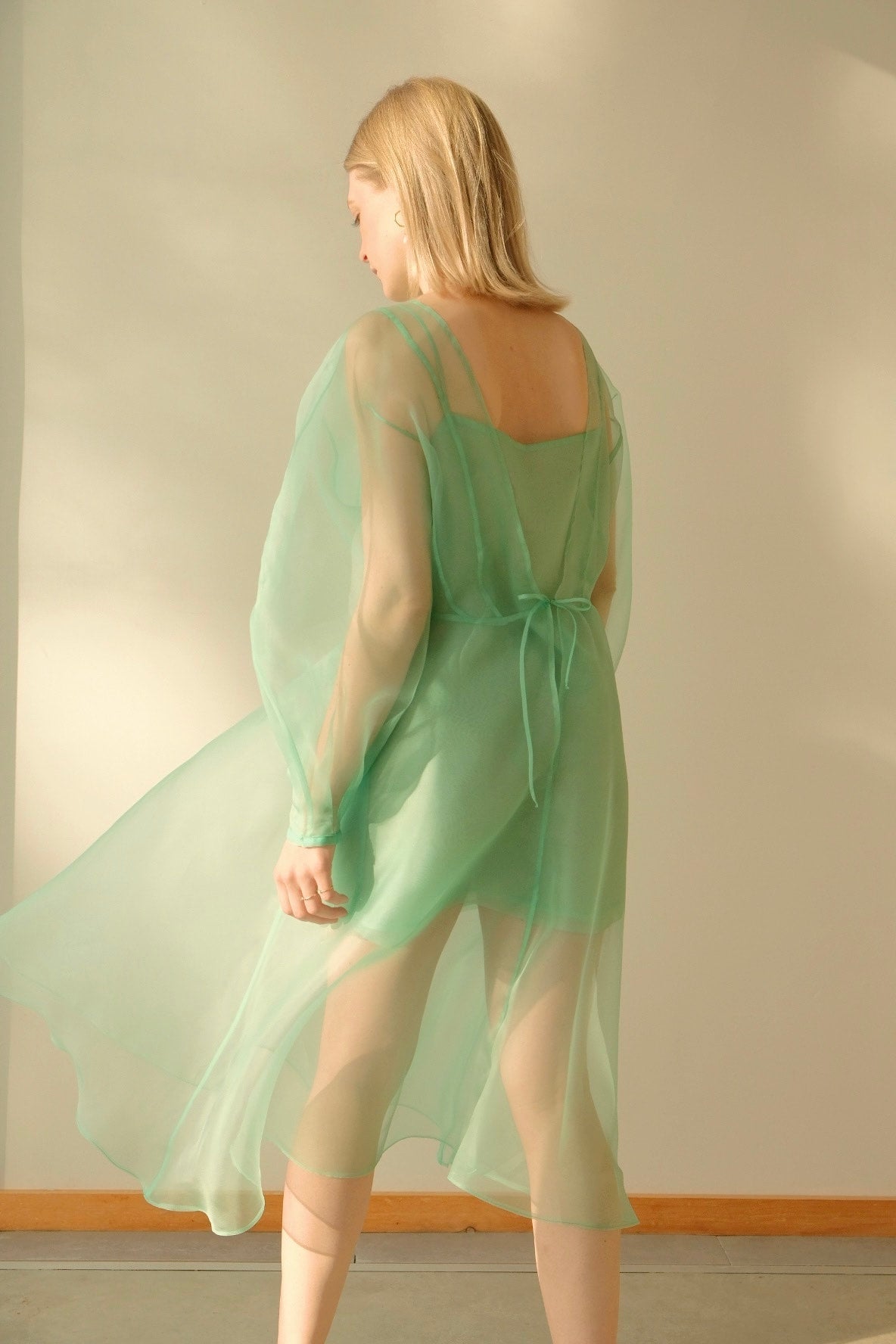 A woman wearing a sheer, mint green Charlotte Dress — Classic by Noémiah stands with her back to the camera. The voluminous cocoon sleeves add an elegant flair as soft lighting casts gentle shadows on her and the surrounding neutral-colored wall.