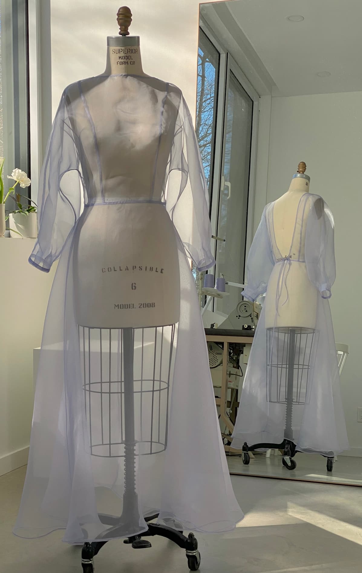 The Noémiah Charlotte Dress — Classic, made from 100% silk organza, is showcased on a form in a sunlit room. Its translucent fabric and voluminous cocoon sleeves create a shimmering reflection in the mirror. The serene setting includes white walls, a window, and a small plant resting on a table.