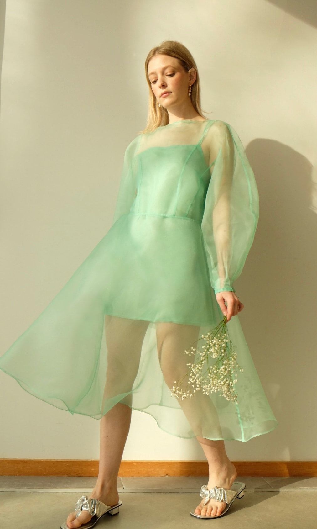 A person adorned in a flowing Charlotte Dress — Classic by Noémiah, made from sheer green 100% silk organza with voluminous cocoon sleeves, stands indoors. They hold a small bouquet of white flowers and wear silver sandals. The softly lit room casts a gentle shadow on the wall behind them.