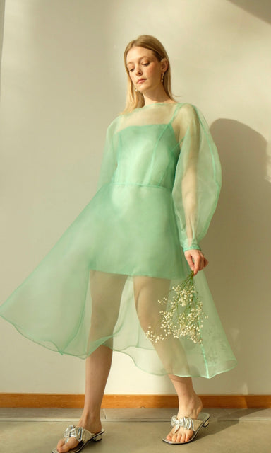 A person adorned in a flowing Charlotte Dress — Classic by Noémiah, made from sheer green 100% silk organza with voluminous cocoon sleeves, stands indoors. They hold a small bouquet of white flowers and wear silver sandals. The softly lit room casts a gentle shadow on the wall behind them.