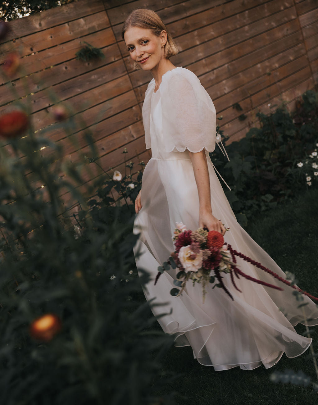 Wearing the Chloé Dress by Noémiah, a woman carries a colorful bouquet as she strolls through a garden. She smiles softly, framed by wooden fencing and lush greenery in the background.