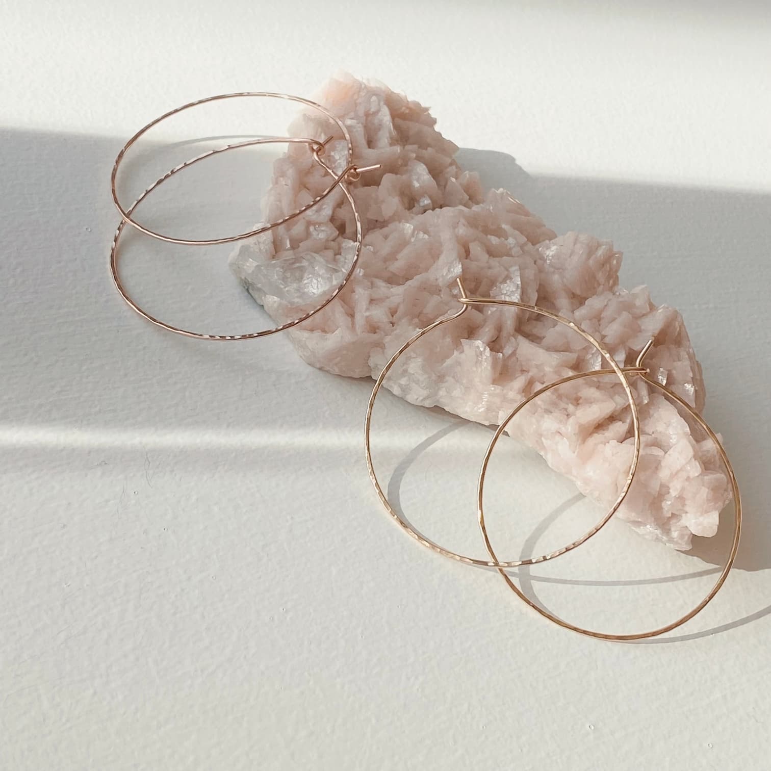 The Clara Earrings by Noémiah, two elegant gold-filled hoops, gently rest on a rough, pale pink rose quartz crystal. Bathed in soft sunlight, they display their subtle hammered texture and cast gentle shadows on the pristine white surface.