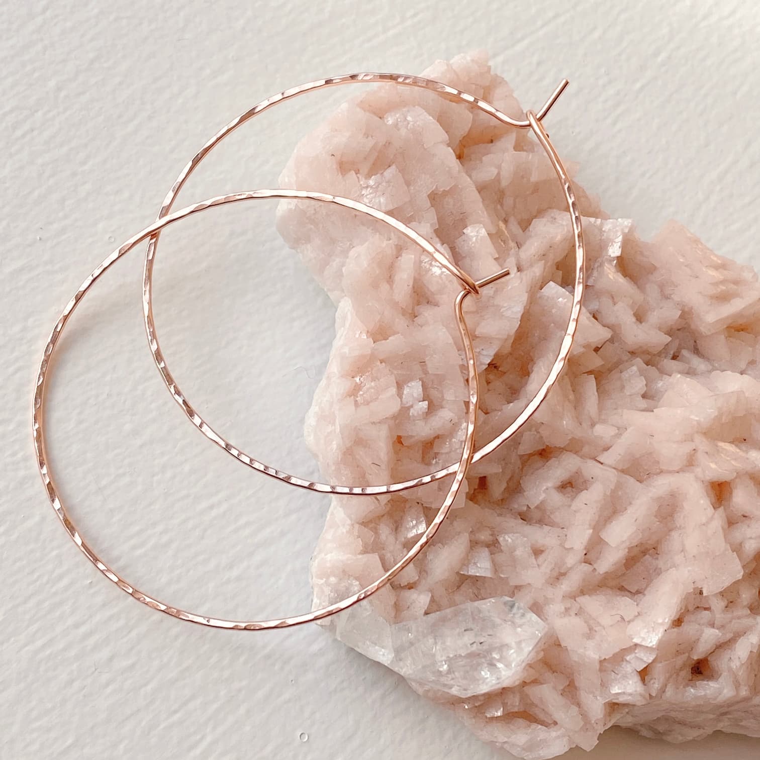 The Clara Earrings by Noémiah, featuring a delicate, hammered texture and gold-filled hoops, are elegantly displayed on a light pink, rough crystalline rock surface against a white background.