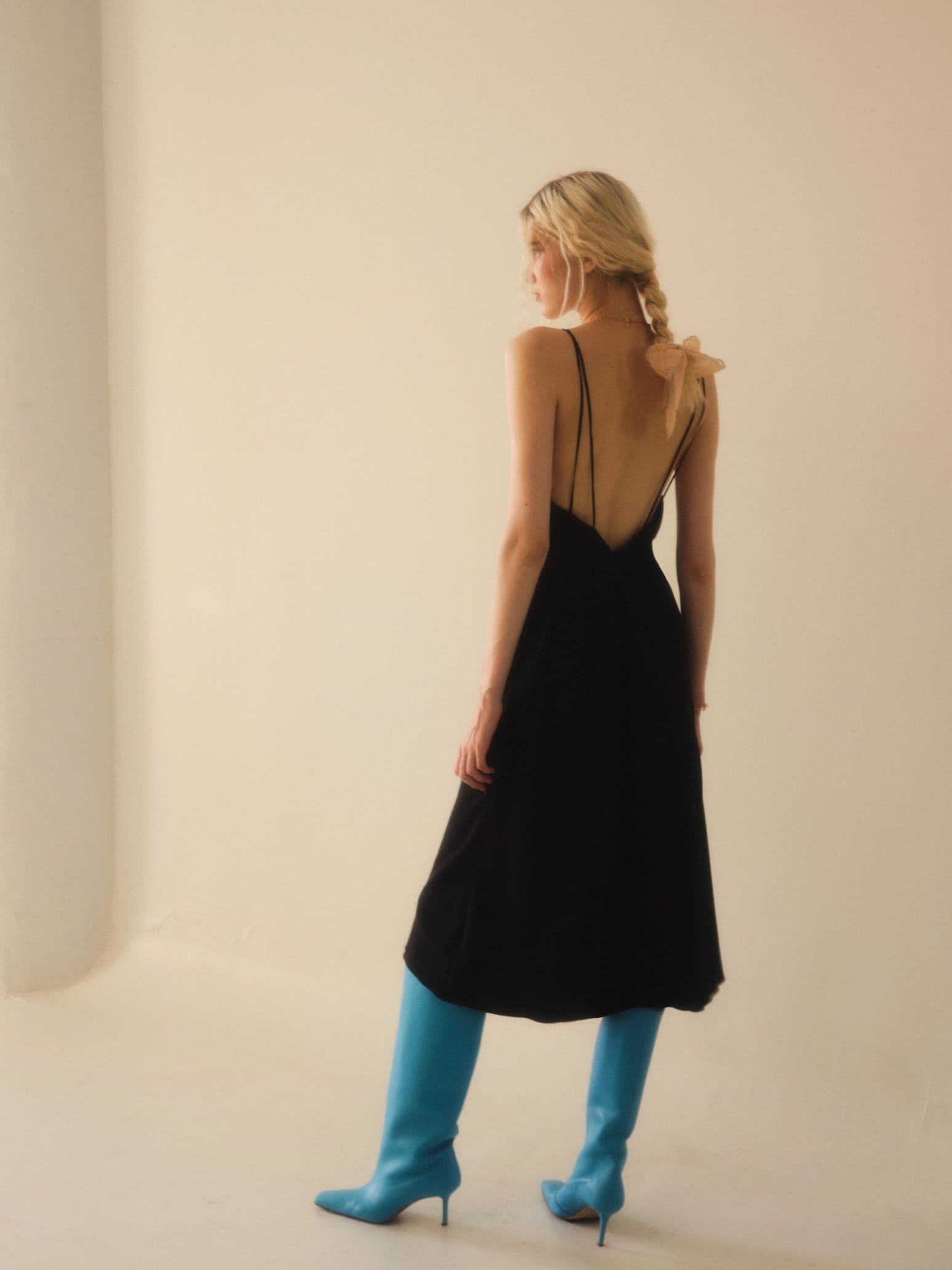 A person with long blonde hair stands in a well-lit room, wearing Noémiah's Claude Dress from the Bridal Edit collection, which features an open back and is paired with knee-high blue boots. The minimalist aesthetic of the setting, along with light-colored walls, enhances their elegant style.