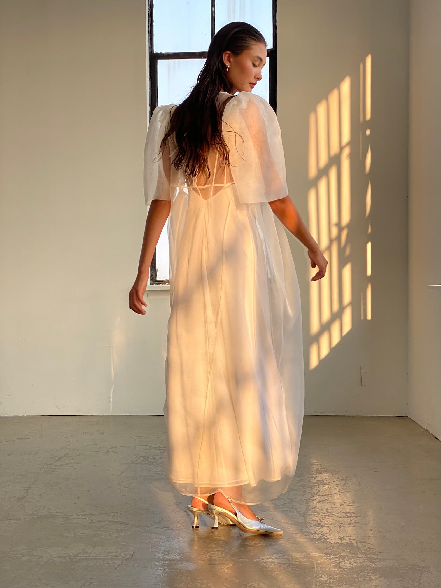 A woman stands in a softly lit room, adorned in Noémiah's Claude Dress — Bridal Edit, a flowing translucent white midi-length piece paired with golden heels. The silk crepe of her bias slip dress catches the light as she gazes slightly away, with sunlight streaming through a tall window and casting warm shadows on her and the wall.