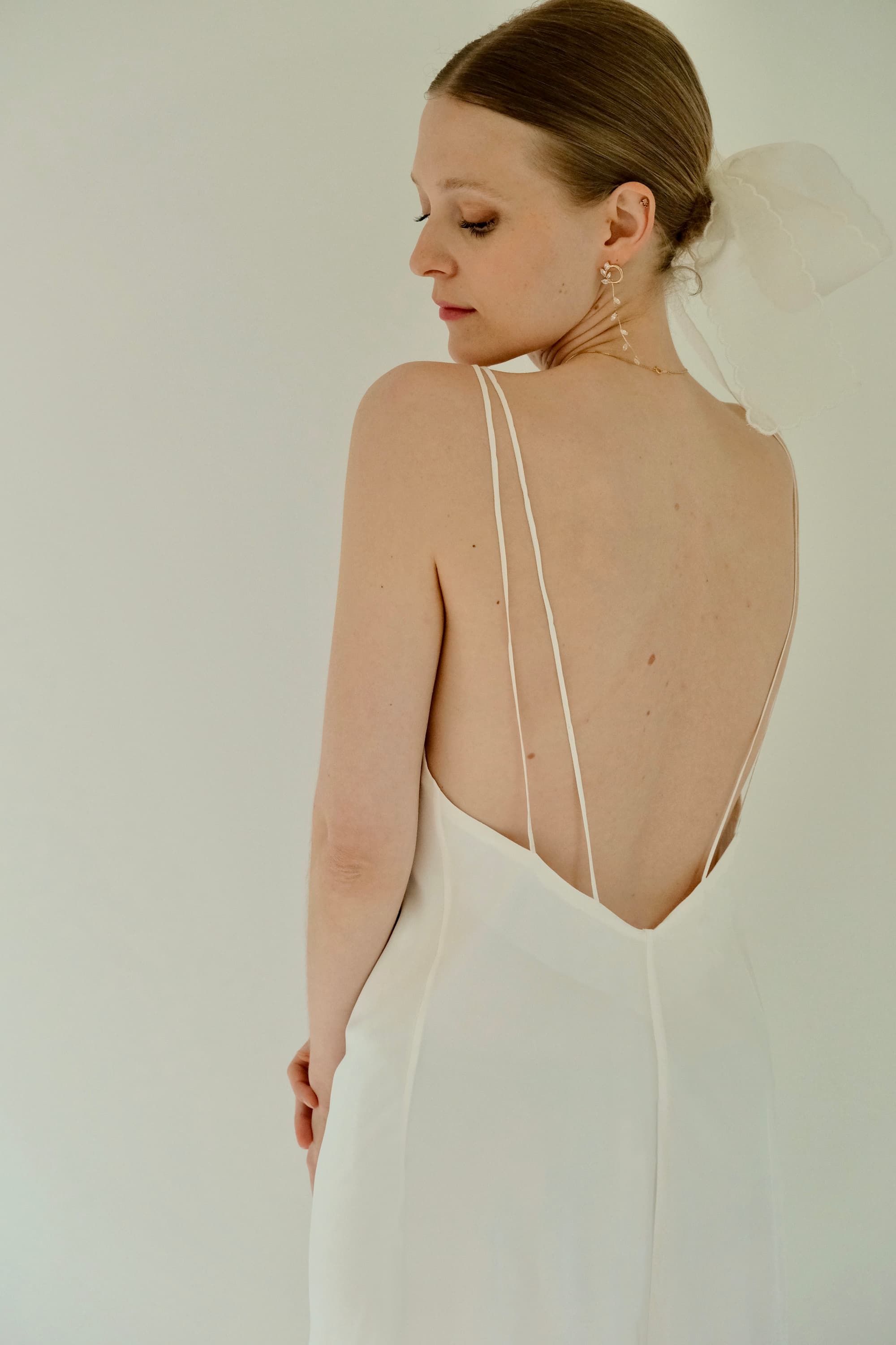 Dressed in the elegant Claude Dress from Noémiah's Bridal Edit, a woman in a chic silk crepe piece cut on the bias at midi length looks over her shoulder. Her hair is styled in a neat bun adorned with a sheer white ribbon, set against a soft, light background.