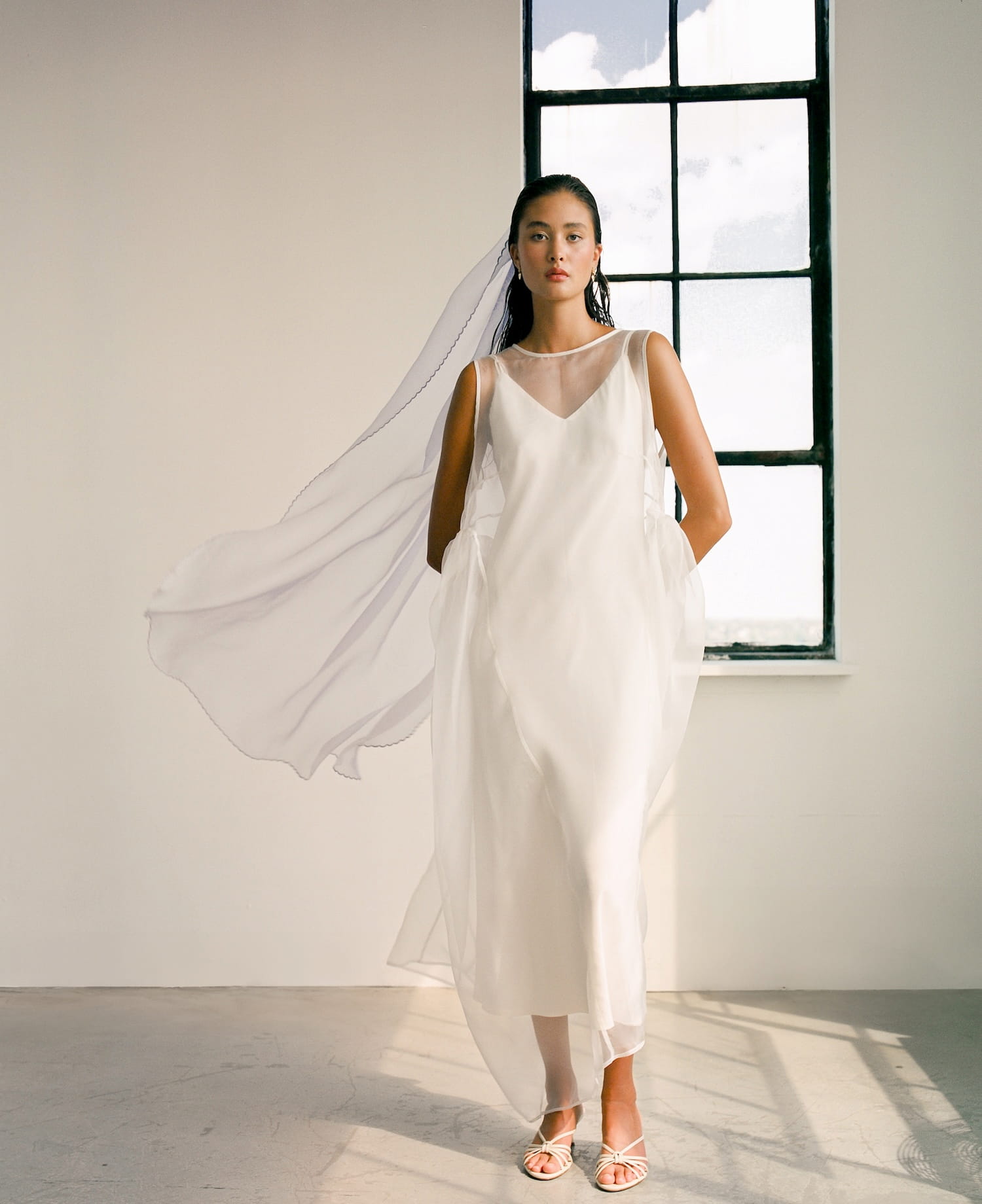 Wearing the Claude Dress — Bridal Edit by Noémiah, a woman in a flowing silk crepe dress and veil gracefully stands in a minimalist room. She is near a tall window with the cloudy sky visible outside, complemented by delicate sandals. The natural light bathes the scene, highlighting the ethereal quality of her midi-length outfit.