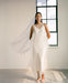 Wearing the Claude Dress — Bridal Edit by Noémiah, a woman in a flowing silk crepe dress and veil gracefully stands in a minimalist room. She is near a tall window with the cloudy sky visible outside, complemented by delicate sandals. The natural light bathes the scene, highlighting the ethereal quality of her midi-length outfit.