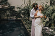 Amidst a backdrop of lush greenery by the pool, a couple savors a joyful moment together. The woman wears the Claude Dress—Bridal Edit by Noémiah, elegantly paired with a headband, while the man sports a white shirt and light pants. Their smiles mirror the vibrant surroundings.