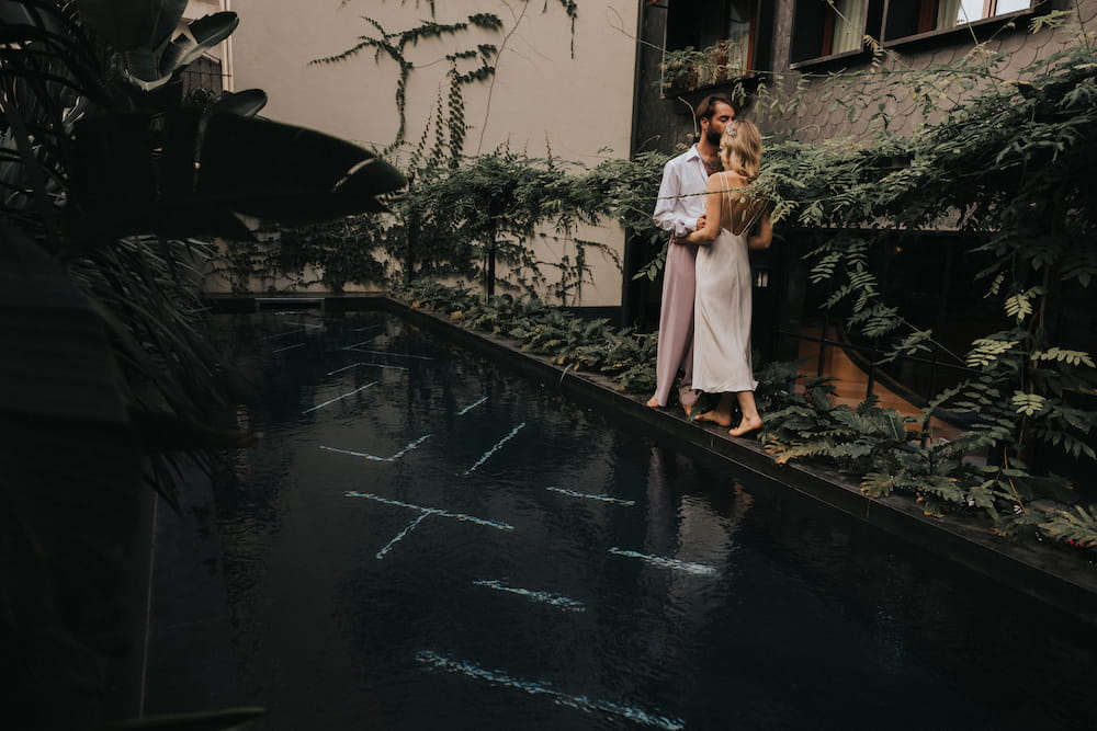 A couple embraces by a dimly lit pool, enveloped by lush greenery. The woman is wearing the elegant "Claude Dress — Bridal Edit" from Noémiah, its silk crepe fabric draping beautifully. The man is dressed in light clothing. The atmosphere feels serene and intimate, with plants climbing the walls of a nearby building.