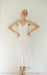 A woman showcases the graceful Claude Dress — Bridal Edit by Noémiah, a minimalist white silk crepe slip dress with thin straps. The elegant bias cut complements her pose against a simple, light backdrop, as she stands with her hands on her hips, accessorized with sandals and a delicate headpiece.