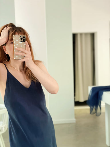 A person in the navy lyocell Claude dress takes a mirror selfie with a smartphone in a modern, minimalist room.