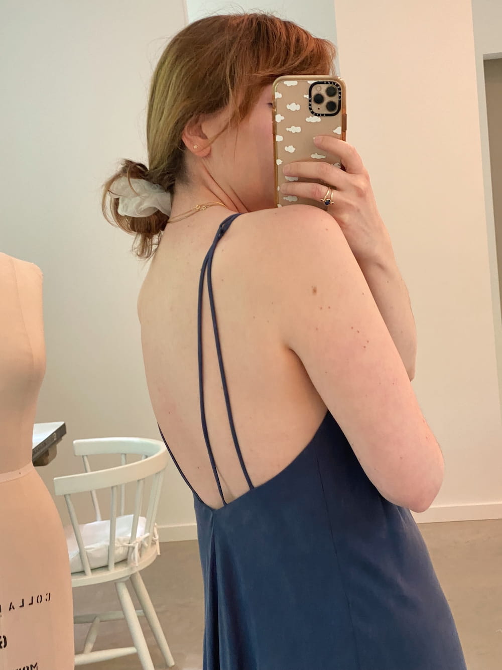 A woman wearing Noémiah's Claude Dress — Lyocell in blue, featuring thin straps and a midi-length design, takes a selfie in front of a mirror. Her hair is styled back with a white scrunchie, and she holds a phone adorned with a cloud-patterned case. In the background, glimpses of a white chair and mannequin highlight the elegance of her lyocell dress.