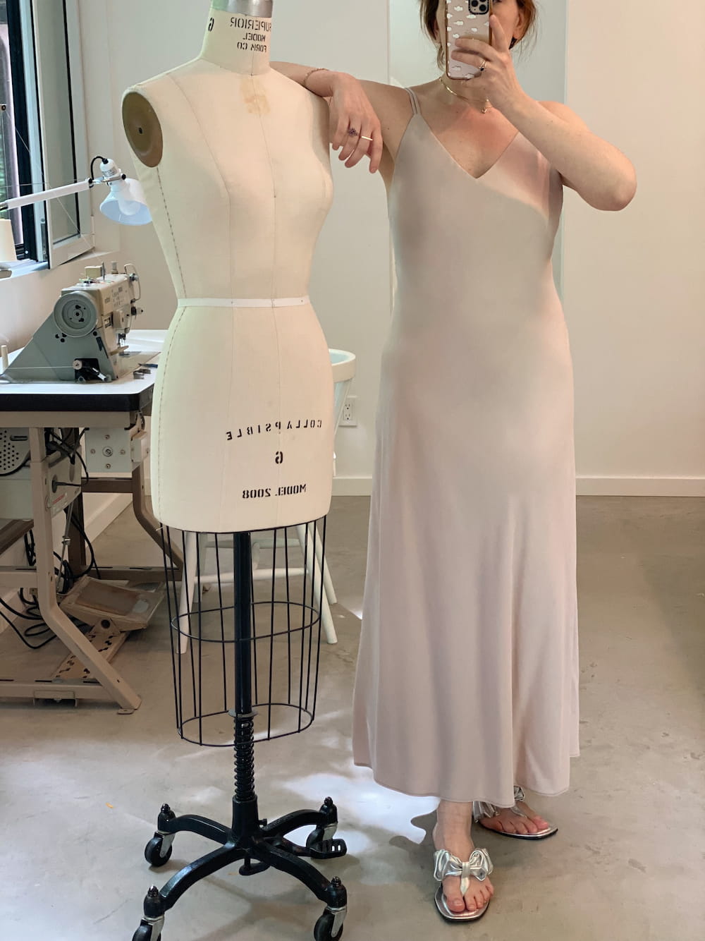 A person takes a mirror selfie in a sewing studio beside a dress form. They are wearing the Claude Dress by Noémiah, which is a long, sleeveless, cream-colored midi-length slip dress crafted from sustainable Lyocell fabric, paired with sandals. In the background, there's a sewing machine and various sewing tools.