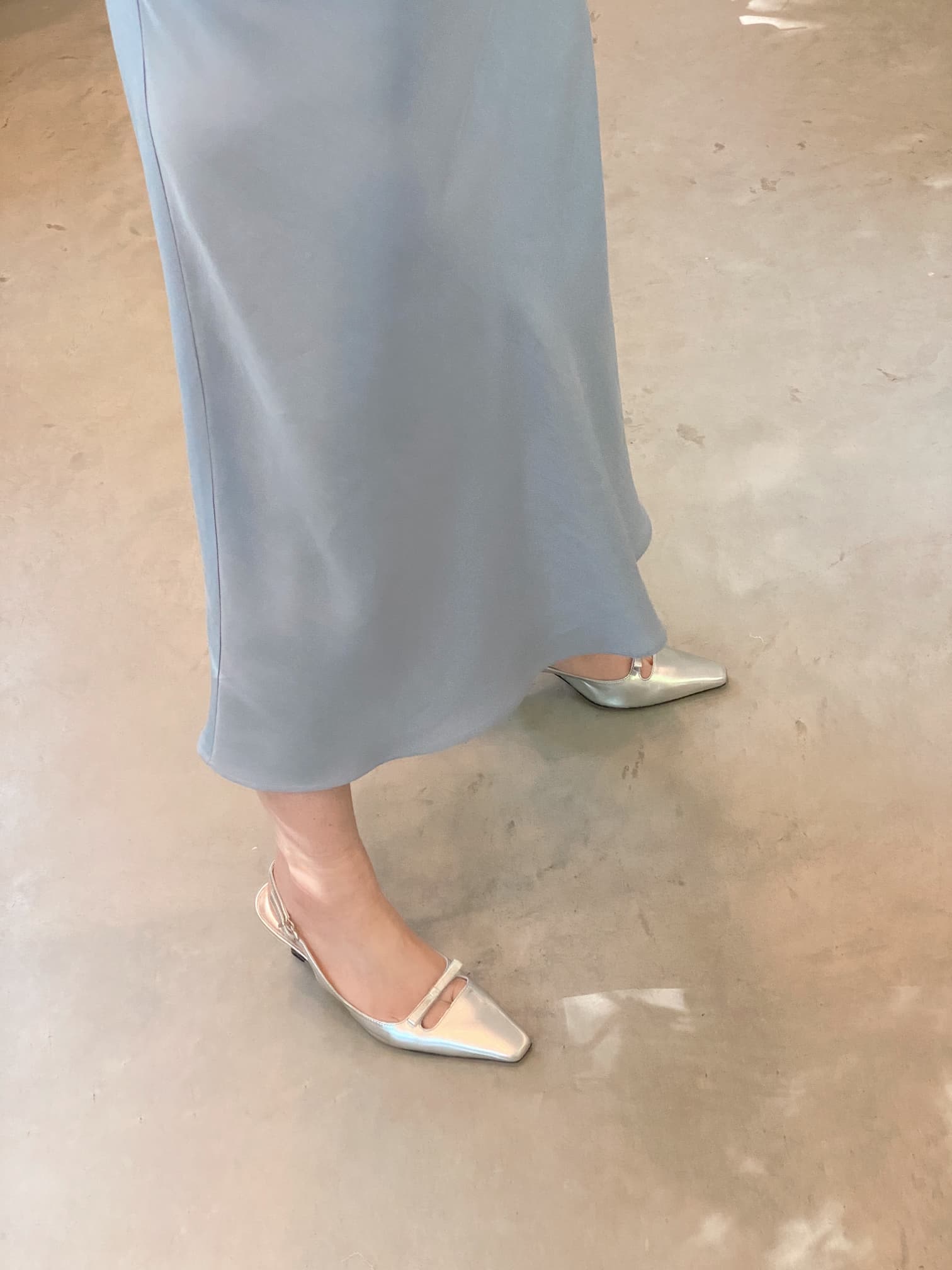 A person wearing a Claude Dress by Noémiah, made from sustainable lyocell in light blue, and silver pointed shoes stands on a gray floor.