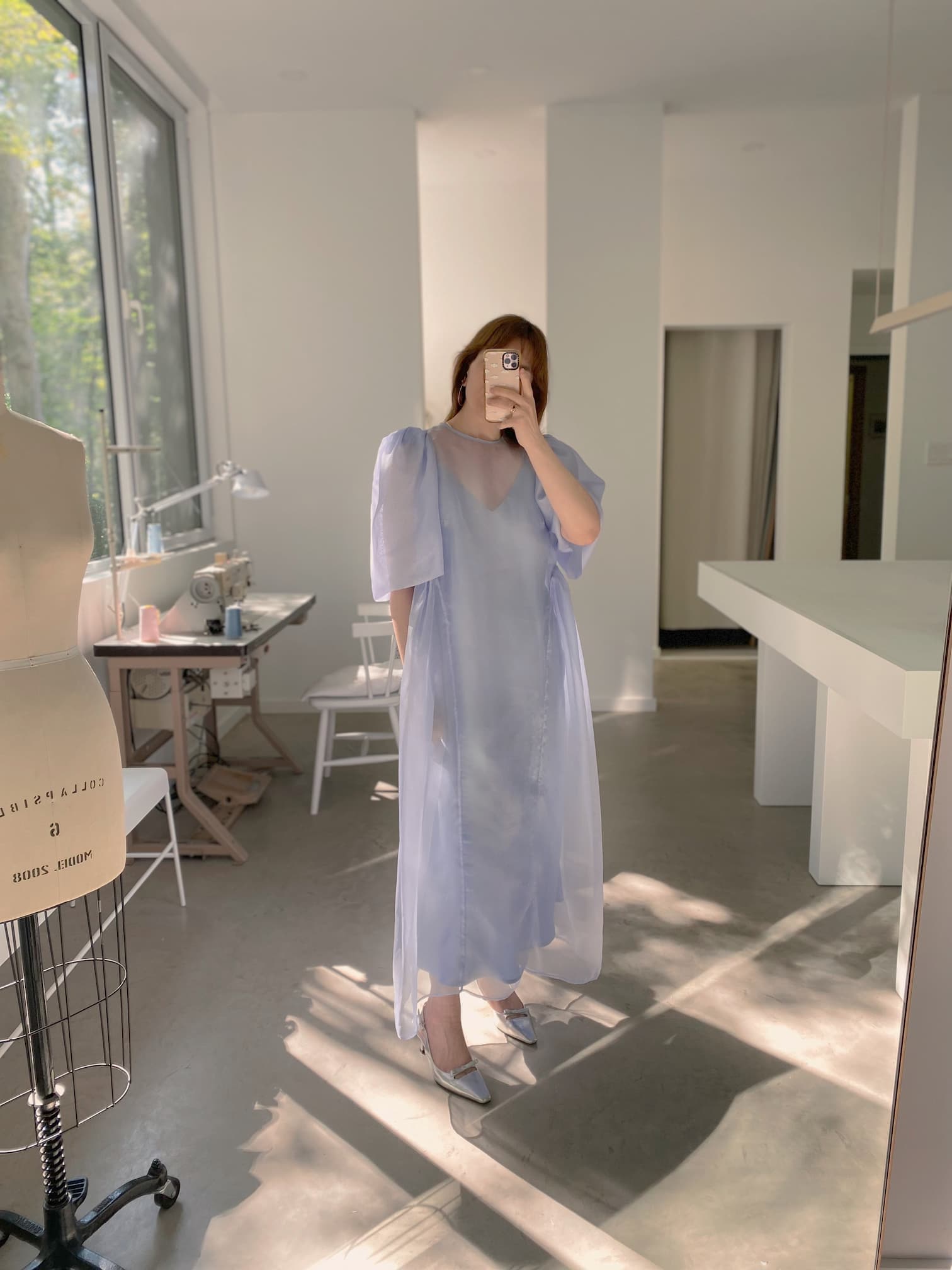 A person wearing the Claude Dress in Lyocell by Noémiah stands in a sunlit room with large windows. The interior includes a sewing mannequin, a table with sewing supplies, and white chairs. The person is holding a phone, taking a mirror selfie.