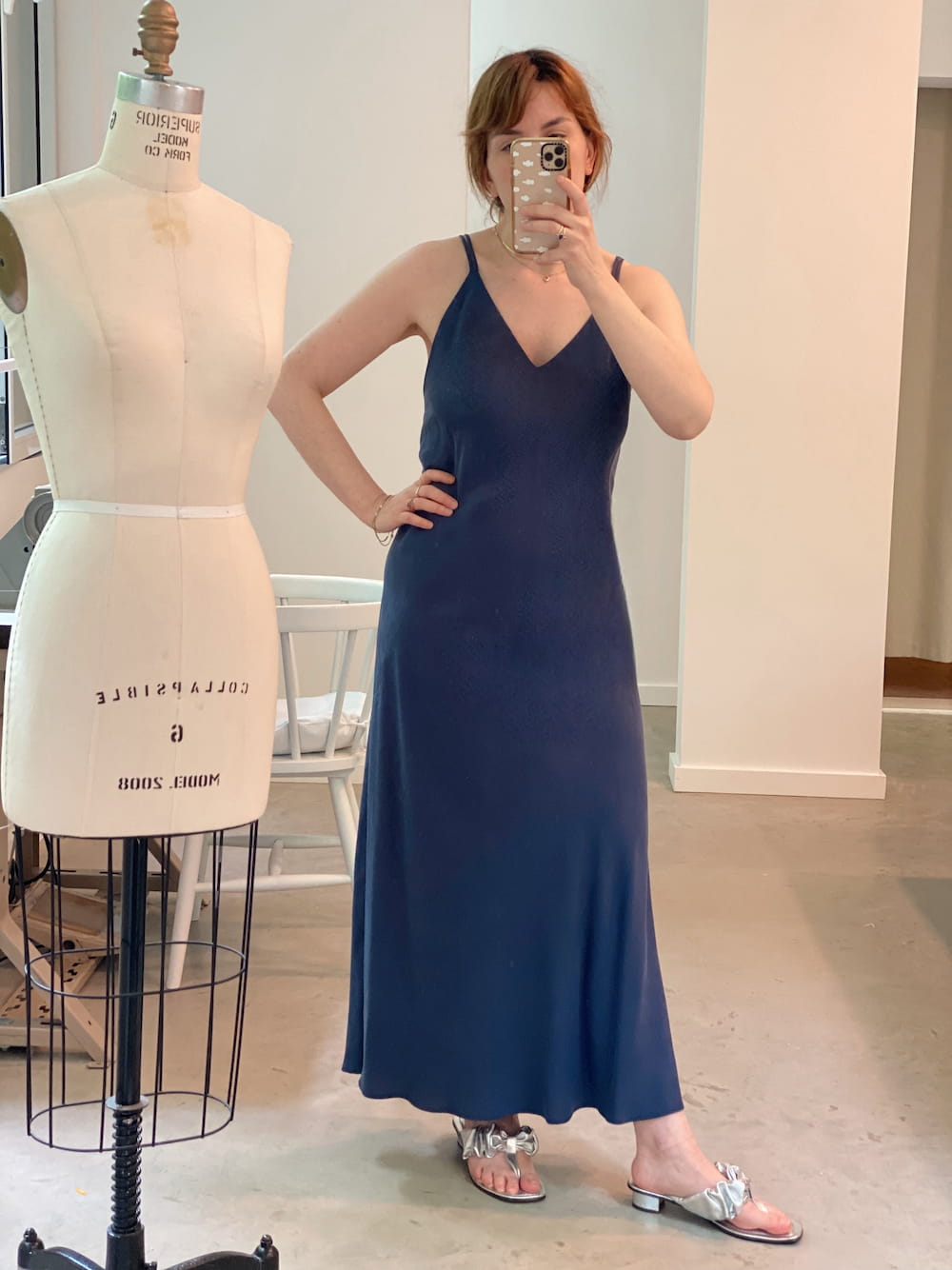 A person in a Claude Dress — Lyocell by Noémiah, which is a blue, midi-length slip dress made from sustainable fabric, takes a mirror selfie in a room with a dress form. They are wearing silver sandals and have short hair.