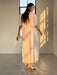 A woman stands in a softly lit room, adorned in Noémiah's Claude Silk Dress — Bridal Edit, a flowing translucent white midi-length piece paired with golden heels. The silk crepe of her bias slip dress catches the light as she gazes slightly away, with sunlight streaming through a tall window and casting warm shadows on her and the wall.