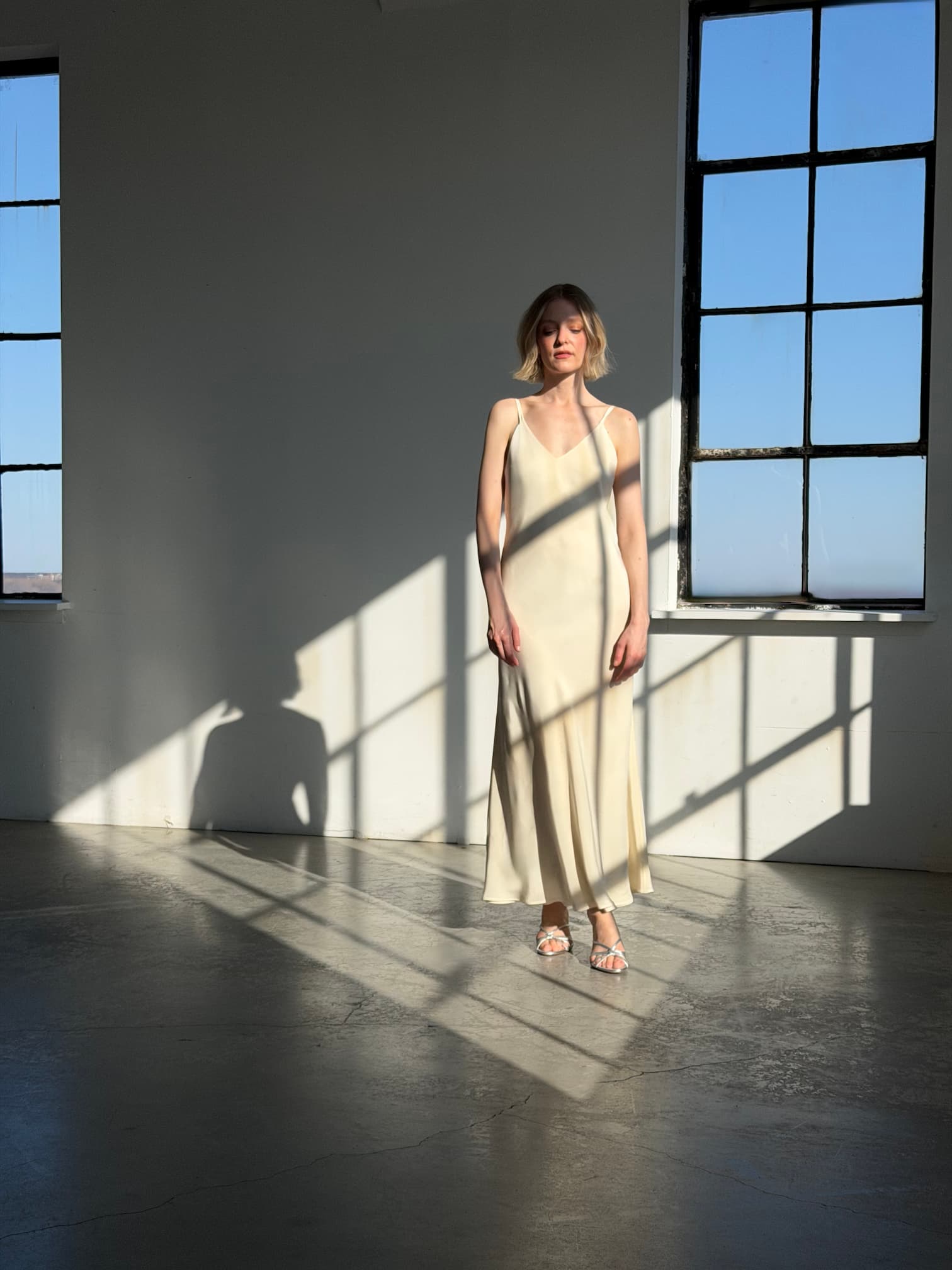 A woman with short blonde hair stands in a sunlit room wearing the Noémiah Claude Silk Dress — Bridal Edit. Light streams through large windows, casting strong shadows on the floor and wall as she looks ahead.