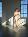 A woman with short blonde hair stands in a sunlit room wearing the Noémiah Claude Silk Dress — Bridal Edit. Light streams through large windows, casting strong shadows on the floor and wall as she looks ahead.
