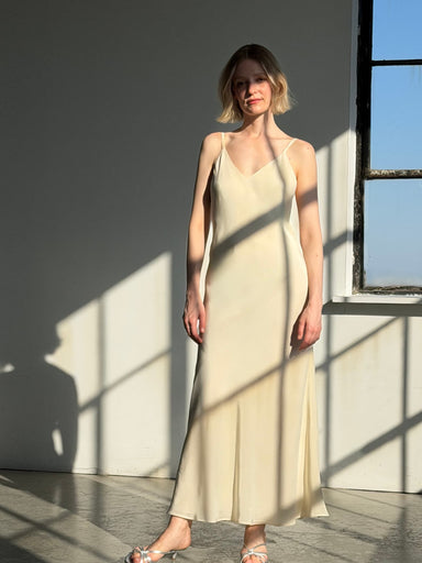 A woman stands barefoot in sunlight by a window, casting shadows on the wall and floor behind her, wearing the Noémiah Claude Silk Dress — Bridal Edit.