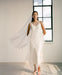 Wearing the Claude Silk Dress — Bridal Edit by Noémiah, a woman in a flowing silk crepe dress and veil gracefully stands in a minimalist room. She is near a tall window with the cloudy sky visible outside, complemented by delicate sandals. The natural light bathes the scene, highlighting the ethereal quality of her midi-length outfit.