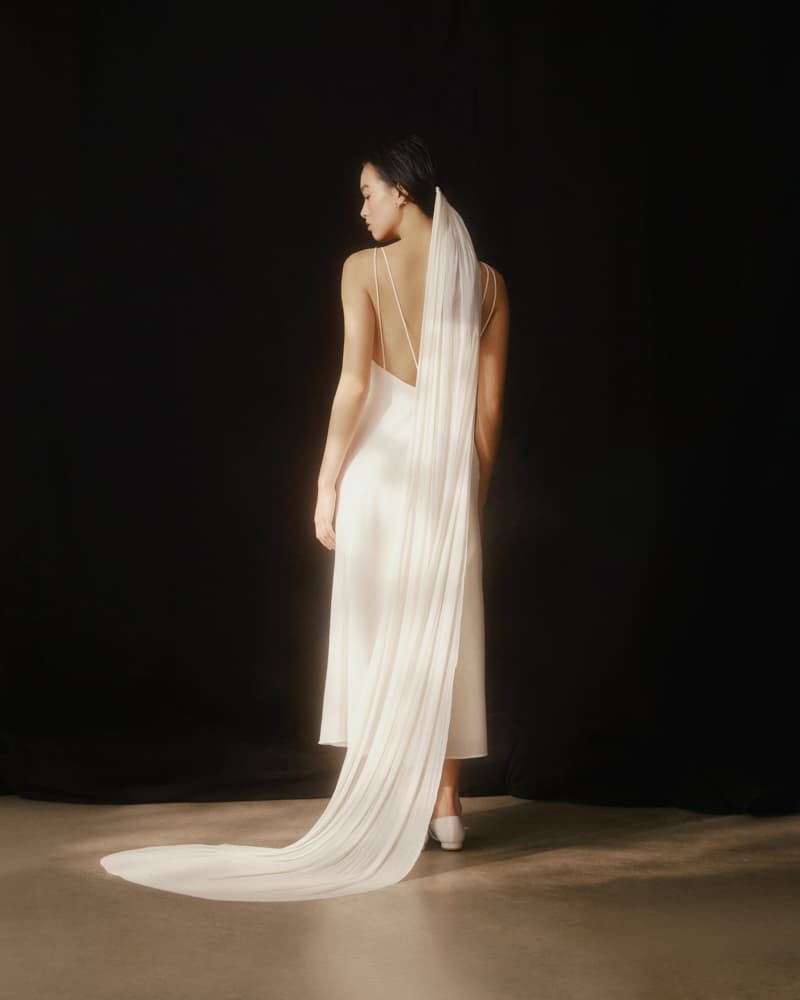 A woman in the Claude Silk Dress — Bridal Edit by Noémiah, featuring a white silk crepe design and long, flowing veil, stands against a dark background. Her back is turned to the camera, with the bias slip design offering a striking contrast between its midi length finish and the backdrop.