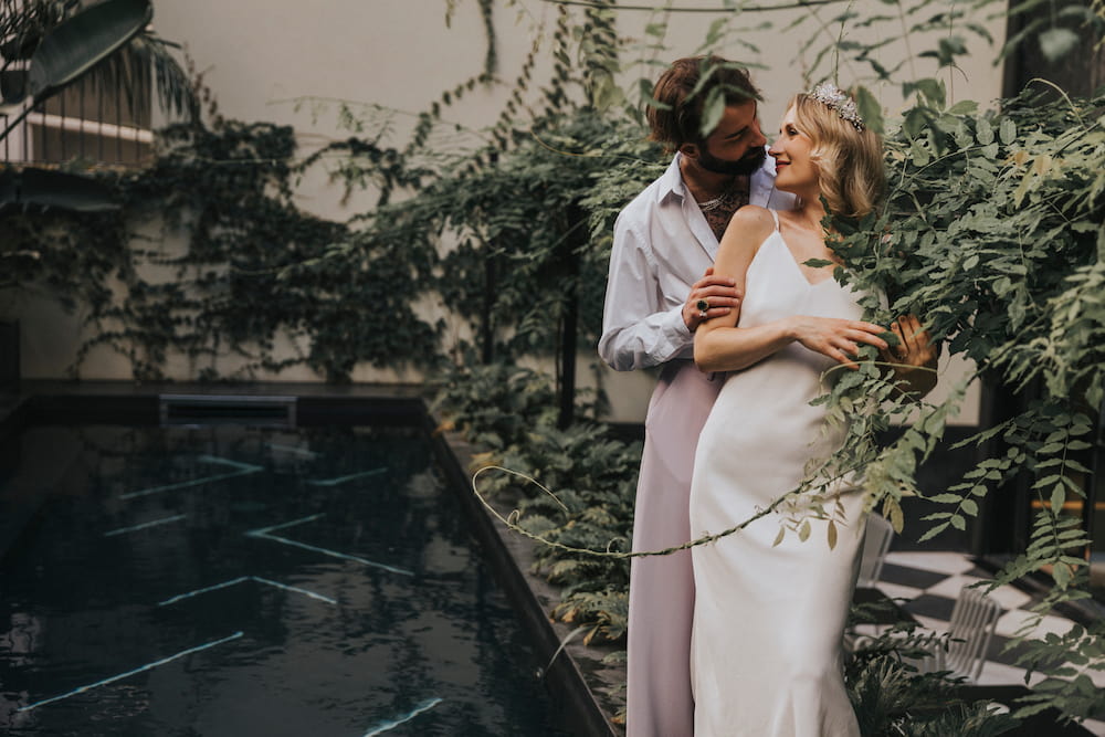 Amidst a backdrop of lush greenery by the pool, a couple savors a joyful moment together. The woman wears the Claude Silk Dress—Bridal Edit by Noémiah, elegantly paired with a headband, while the man sports a white shirt and light pants. Their smiles mirror the vibrant surroundings.