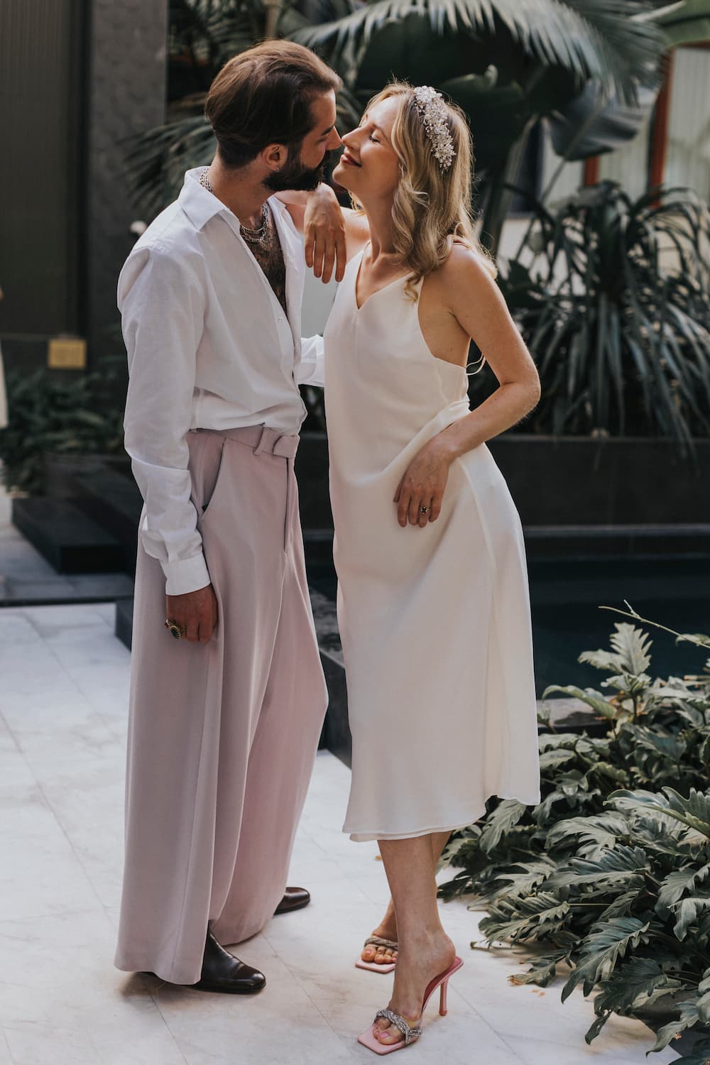 In a stylish indoor setting surrounded by lush greenery, a couple stands close together. The man is dressed in a white shirt and wide pants, while the woman captivates in Noémiah's Claude Silk Dress — Bridal Edit and elegant heels. They lean in towards each other, smiling affectionately.