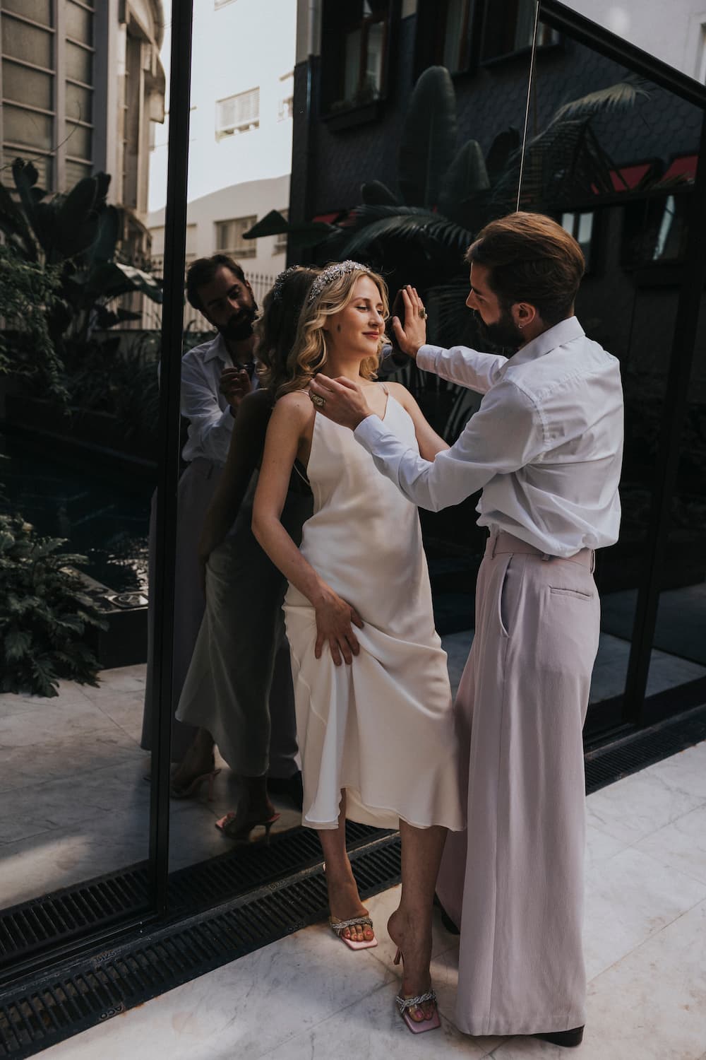 A woman in the Claude Silk Dress — Bridal Edit by Noémiah and a man in a white shirt with light pants stand close together in front of a large glass window, with greenery reflecting behind them. The woman smiles, her eyes closed, as the man gently holds her shoulders.