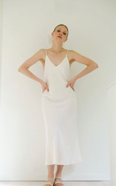 A woman showcases the graceful Claude Silk Dress — Bridal Edit by Noémiah, a minimalist white silk crepe slip dress with thin straps. The elegant bias cut complements her pose against a simple, light backdrop, as she stands with her hands on her hips, accessorized with sandals and a delicate headpiece.