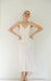 A woman showcases the graceful Claude Silk Dress — Bridal Edit by Noémiah, a minimalist white silk crepe slip dress with thin straps. The elegant bias cut complements her pose against a simple, light backdrop, as she stands with her hands on her hips, accessorized with sandals and a delicate headpiece.