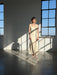 A woman with short blonde hair stands in a sunlit room wearing the Noémiah Claude Silk Dress — Bridal Edit. Light streams through large windows, casting strong shadows on the floor and wall as she looks ahead.