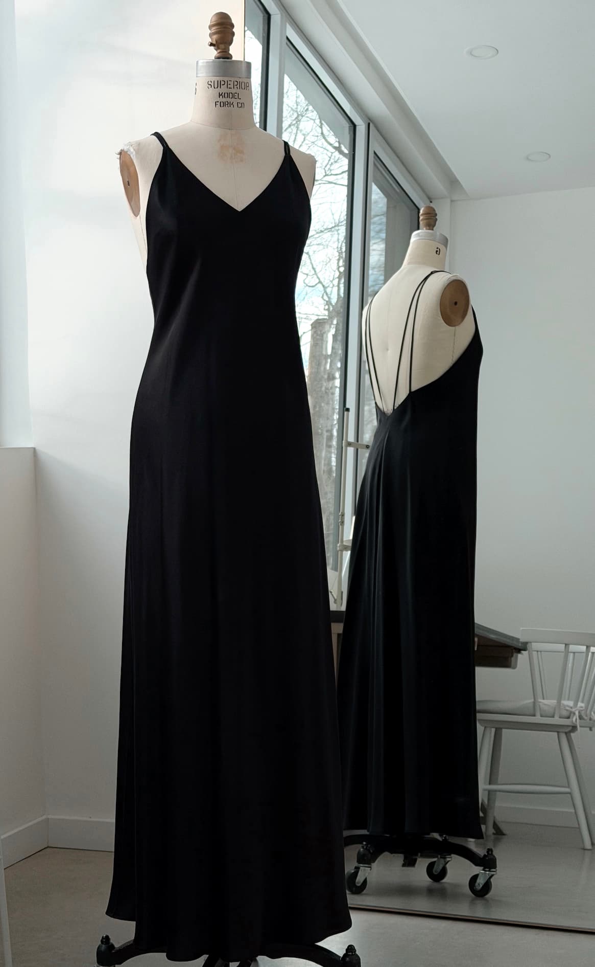 The Claude Silk Satin Dress — Bridal Edit by Noémiah, a long black slip dress with thin straps, is displayed on a dress form with its front and back reflected in a large mirror by the window of a bright, modern room.