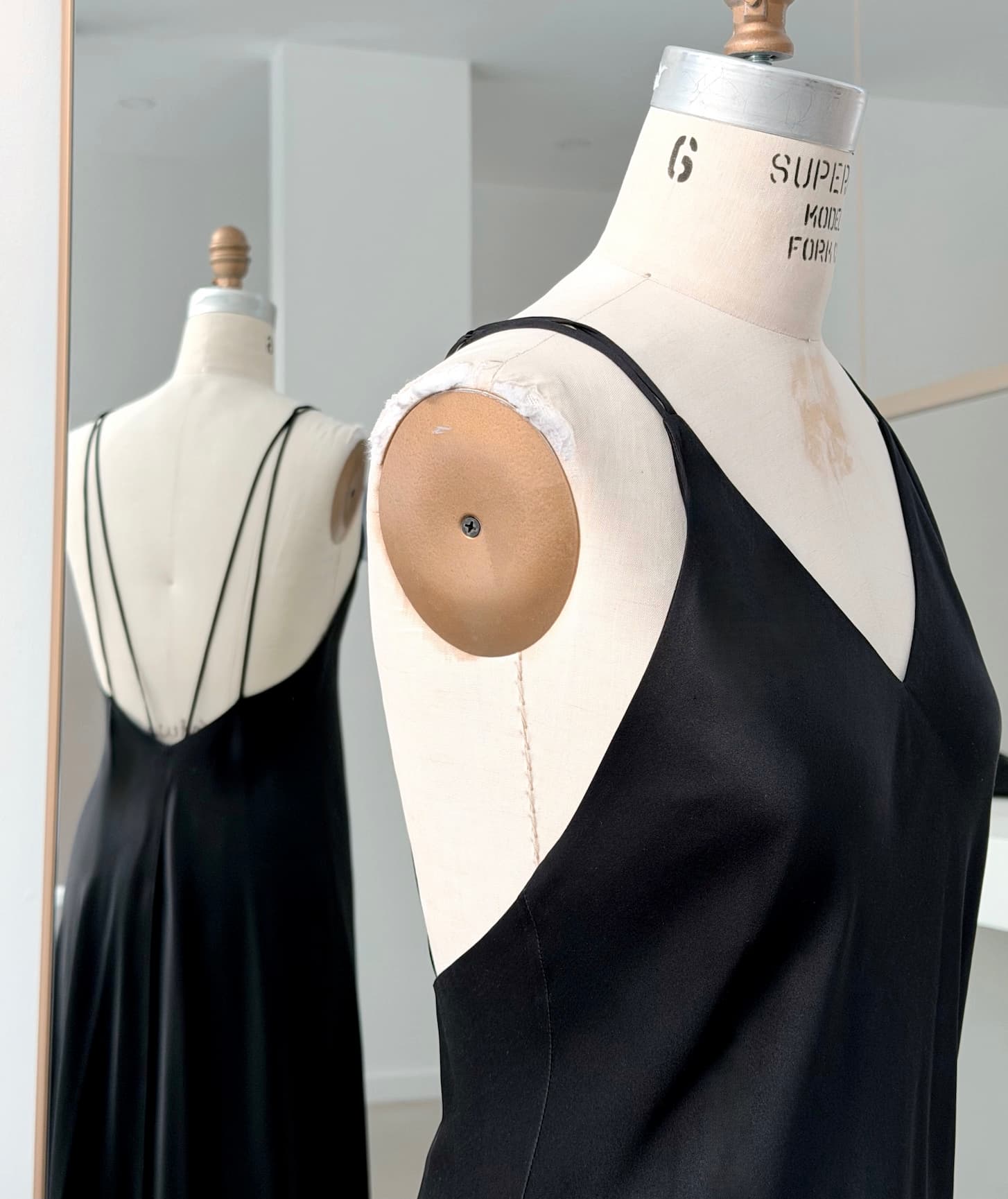 A dress form showcases the Noémiah Claude Silk Satin Dress — Bridal Edit, featuring thin straps and a low back, reflected in a mirror within a bright, minimalistic room.