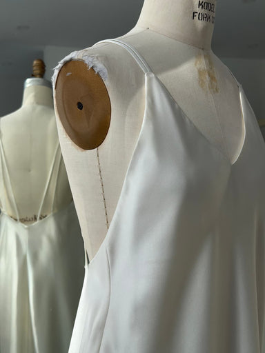 The Claude Silk Satin Dress — Bridal Edit by Noémiah, a white slip dress with thin straps, is displayed on a dress form. The softly lit scene shows the frayed mannequin shoulder and the dress’s back reflected in a mirror.