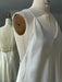 The Claude Silk Satin Dress — Bridal Edit by Noémiah, a white slip dress with thin straps, is displayed on a dress form. The softly lit scene shows the frayed mannequin shoulder and the dress’s back reflected in a mirror.