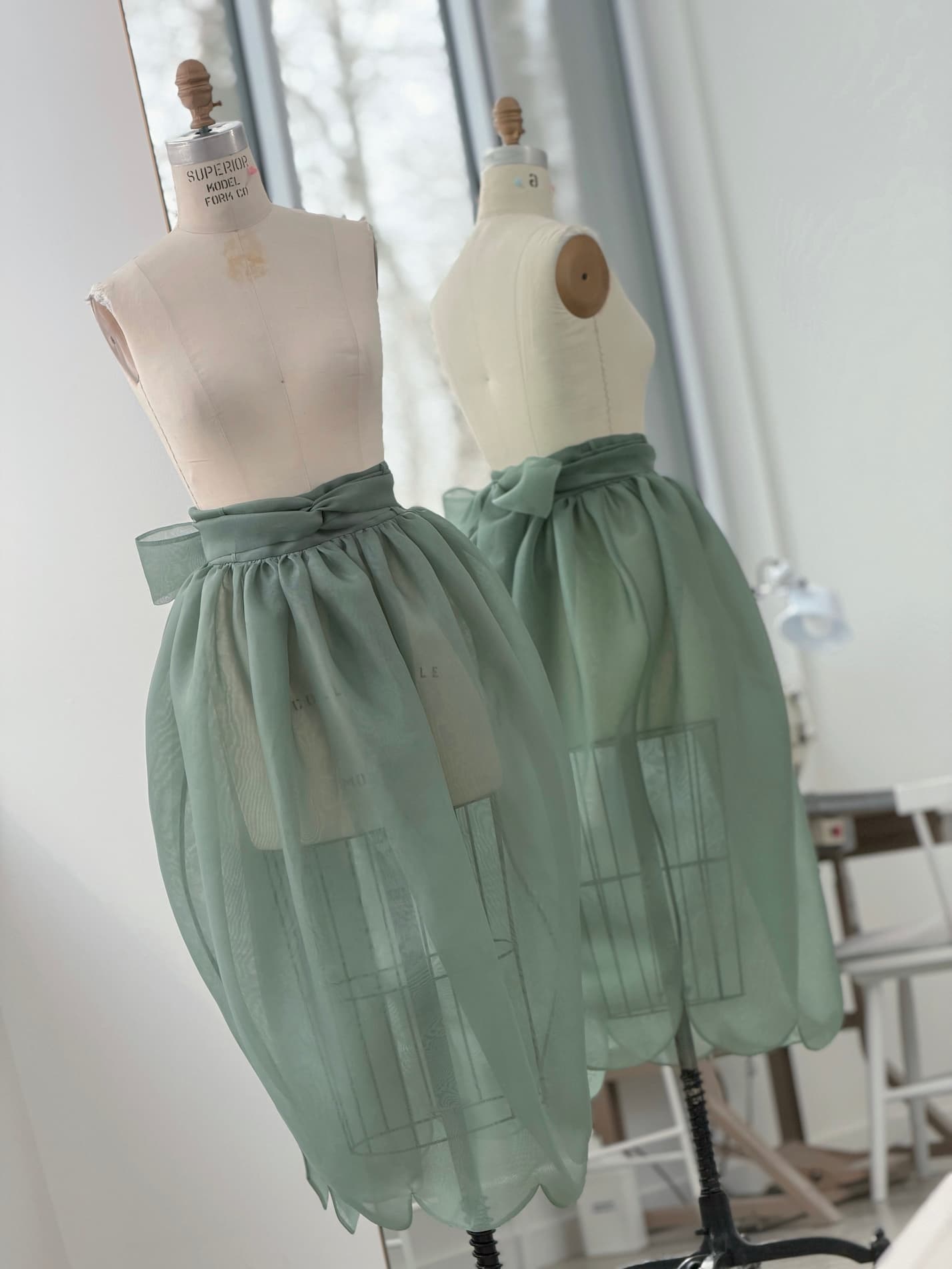 A dress form displays the Clémentine Skirt by Noémiah—a sheer mint green silk organza piece with a waist bow—standing beside a mirror in a bright studio with large windows and sewing equipment in the background.