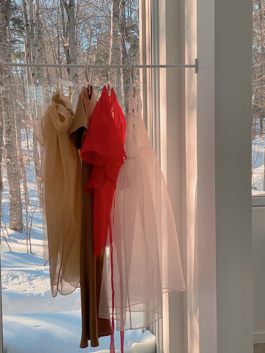 Three elegant dresses, including a Florence Silk Dress—Classic by Noémiah, hang on a rack by a sunlit window. The beige, red, and white garments are made of sheer silk organza. A snowy forest scene is visible through the glass outside.