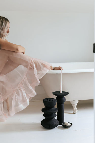 A woman wearing the Coco Ruffle Hemline Silk Dress by Noémiah sits on the edge of a white clawfoot bathtub. Beside the tub are black decorative items, including a tall candlestick with a pink candle. The room has a minimal and bright aesthetic.