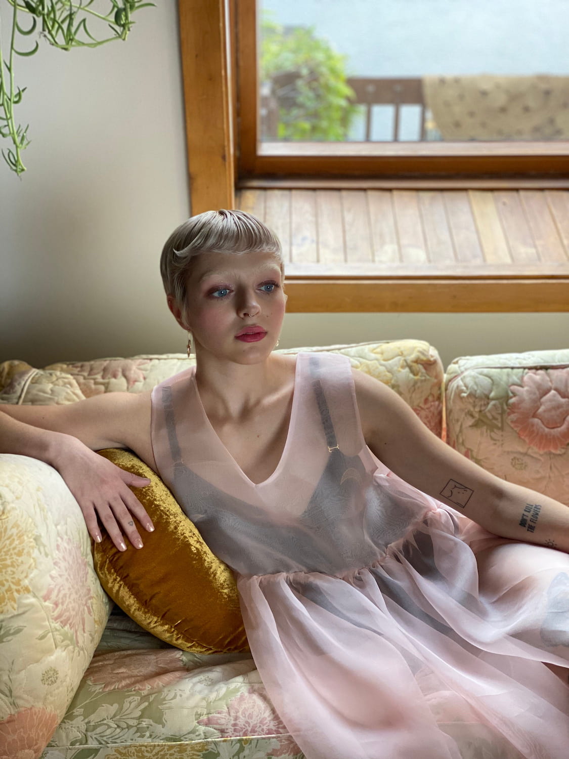 A person with short hair is sitting on a floral couch, wearing the sheer, light pink Coco Silk Dress by Noémiah. They have a focused expression and one arm resting on a yellow pillow. The background shows a window with greenery outside.