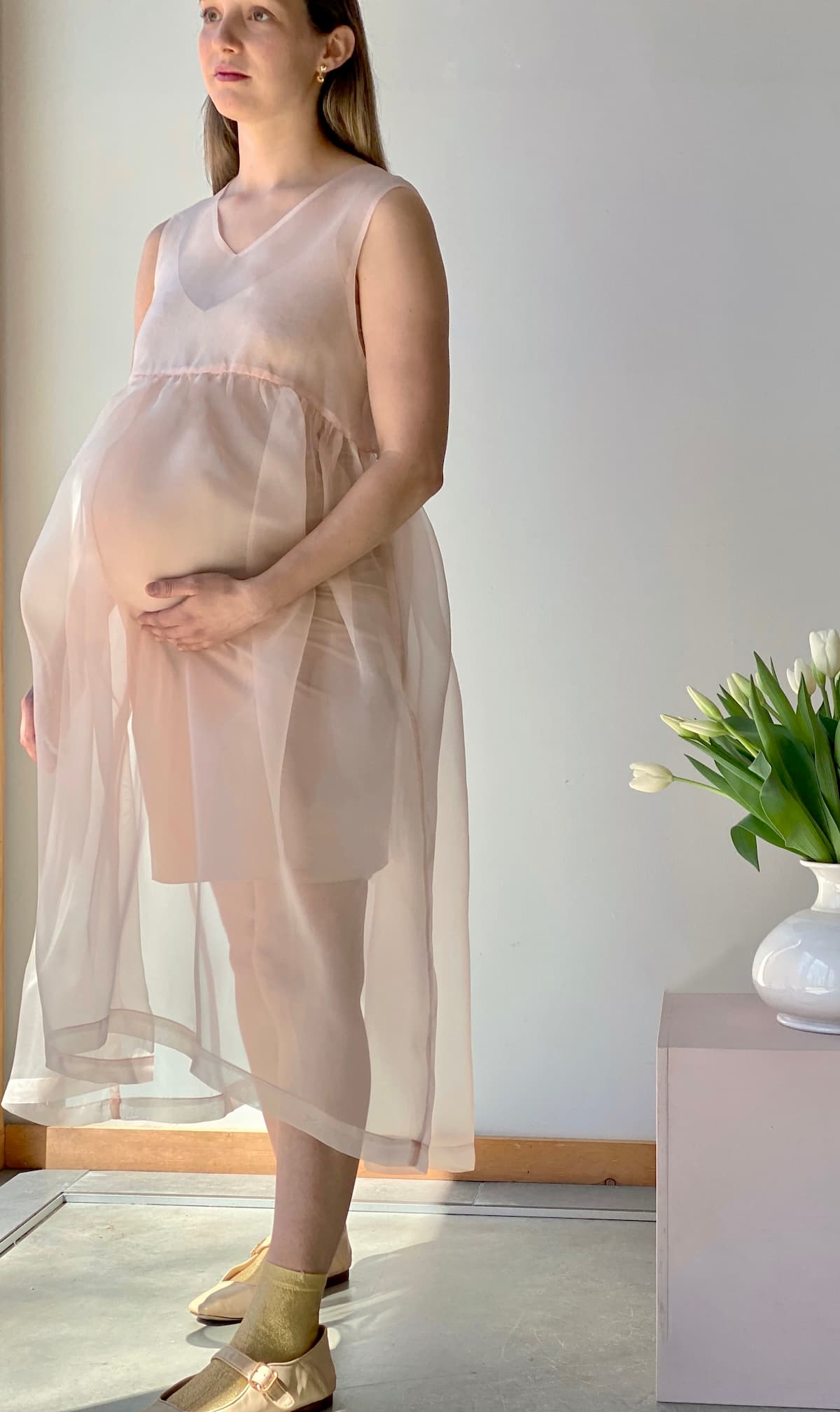 A pregnant woman stands in a well-lit room, wearing a sheer pink Coco Silk Dress by Noémiah and gold shoes. Her light brown hair cascades as she looks ahead. Beside her, a small table showcases a white vase filled with white tulips, adding to the serene scene with its relaxed fit charm.