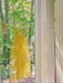 A Coco Silk Dress by Noémiah, crafted from sheer yellow silk organza, hangs on a clothesline indoors near a large window. Sunlight filters through the trees outside, creating soft shadows on the floor and walls and accentuating its relaxed fit.